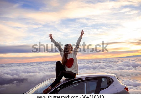 Young woman in sweater with heart shape rising hands sitting on the car roof above the clouds on the sunrise. Enjoying beautiful cloudscape in the morning