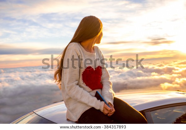 Young woman in sweater with heart shape enjoying\
beautiful cloudscape sitting on the car roof above the clouds on\
the sunrise