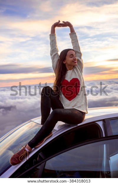Young woman in sweater with heart shape\
rising hands sitting on the car roof above the clouds on the\
sunrise. Enjoying beautiful cloudscape in the\
morning