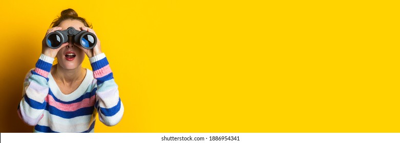 Young woman in sweater looking through binoculars on yellow background.