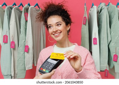 Young Woman In Sweater Stand Near Clothes Rack With Tag Sale In Store Showroom Hold Wireless Modern Bank Payment Terminal Process Acquire Credit Card Payments Isolated On Plain Pink Background Studio