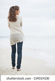 Young Woman In Sweater Walking On Lonely Beach . Rear View