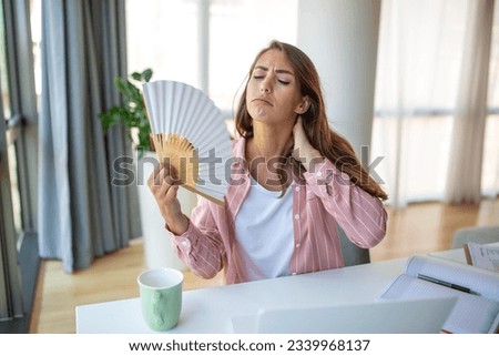 A young woman sweats profusely while working on her laptop in an overheated home office. With no air conditioner, she clings to a wave fan, battling the intense summer heat.