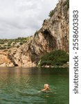 A young woman swims in the rocky, mineral-rich therapeutic waters of Lake Vouliagmeni on an autumn day in Greece. Vertical photo. 