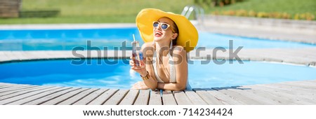 Young woman in swimsuit with big yellow sunhat relaxing with a bottle of fresh drink sitting on the poolside outdoors. Panoramic cropped image
