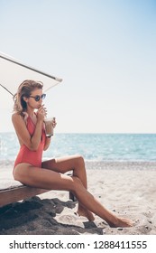 Young Woman In Swimsuit With Cocktail Glass On White Beach Sitting On Sunbed