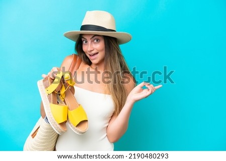 Young woman in swimsuit holding summer sandals isolated on blue background surprised and pointing finger to the side