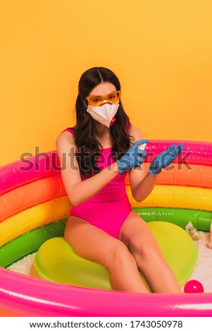 young woman in swimsuit, sunglasses, latex gloves and medical mask sitting in inflatable pool and applying sanitizer on yellow