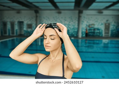 A young woman in a swimsuit and swim cap holding her head in front of an indoor swimming pool. - Powered by Shutterstock