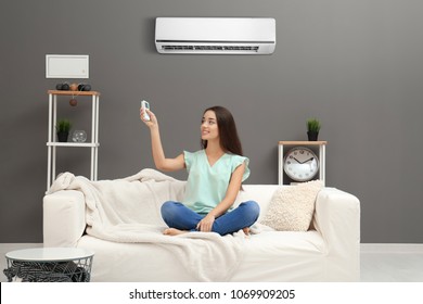 Young woman switching on air conditioner while sitting on sofa at home - Shutterstock ID 1069909205