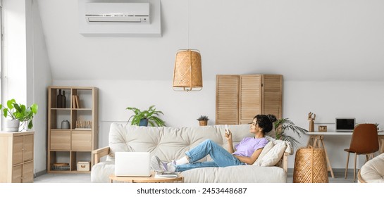 Young woman switching on air conditioner while sitting on sofa at home - Powered by Shutterstock