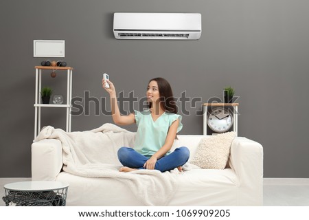 Young woman switching on air conditioner while sitting on sofa at home
