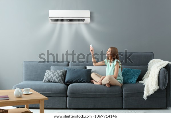 Young woman switching on air conditioner while\
sitting on sofa at home