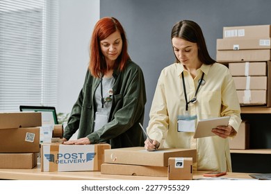 Young woman with tablet writing down address of receiver on packed box while standing next to colleague sealing parcels with cellotape