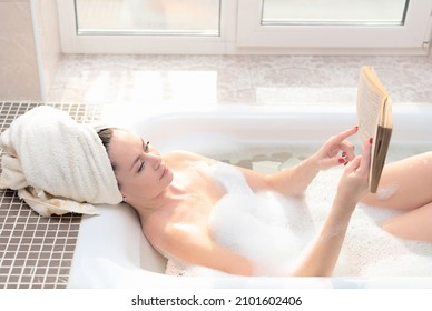 Young woman takes a bath with lush foam near the window reading the book. Personal hygiene, healthcare, relax at home concept - Powered by Shutterstock