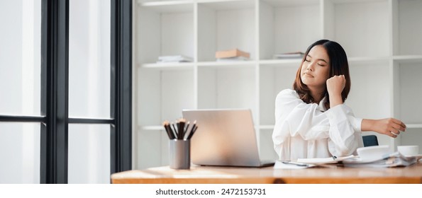 A young woman takes a break from work, stretching her arms at a home office desk with a laptop and coffee, in a bright modern workspace. - Powered by Shutterstock