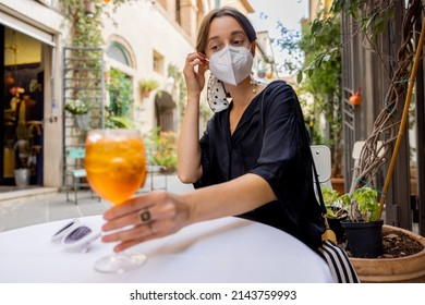 Young Woman Takes Off Face Mask While Sitting And Drinking Spritz Aperol At Italian Restaurant Outdoors. Concept Of Social Rules During Pandemic And Travel Italy