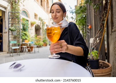 Young Woman Takes Off Face Mask While Sitting And Drinking Spritz Aperol At Italian Restaurant Outdoors. Concept Of Social Rules During Pandemic And Travel Italy