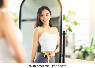  young woman takes a paper measuring ruler and measures her waist size before mirror. She is very satisfied with the results of the weight loss and the current figure. - Shutterstock ID 1989700694