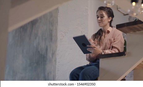 Young woman takes a rest with digital tablet computer. Stock footage. Close-up of beautiful smiling woman in pink shirt and jeans sitting on the wooden stairs at home and typing on the tablet screen.