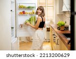 Young woman takes some food from a fridge filled with healthy vegetables and fruits while cooking at home. Concept of healthy lifestyle and eating