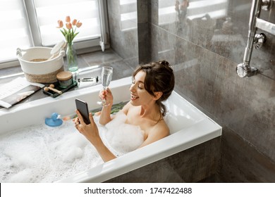 Young woman taking a bath, drinking sparkling wine, talking on phone, lying in bathtub with foam near the window at home - Powered by Shutterstock