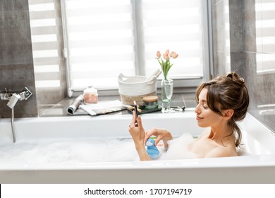 Young woman taking a bath, lying with a smart phone in bathtub with foam and rubber duck in the bathroom at home - Powered by Shutterstock