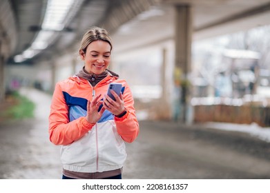 Young Woman Taking Break From Jogging In The City During Winter And Using Smartphone. Winter Fitness And Technology.