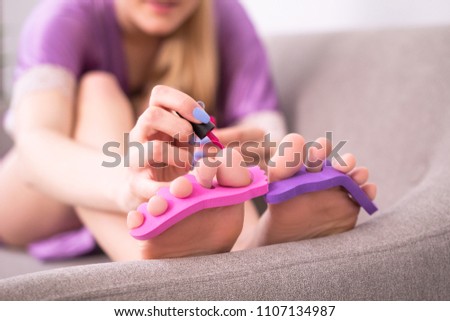 Young woman taking care of her nails on sofa in living room. Blondhaired girl doing her pedicure at home on couch with toe separators in her feet.