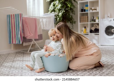 Young Woman Taking Care Of Sweet Blonde Daughter, Little Girl Helps Woman Sort Clothes For Laundry, Baby Sits In Bowl, Mom Hugs Baby.