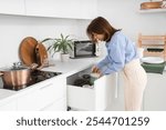 Young woman taking cooking pot from open drawer in kitchen