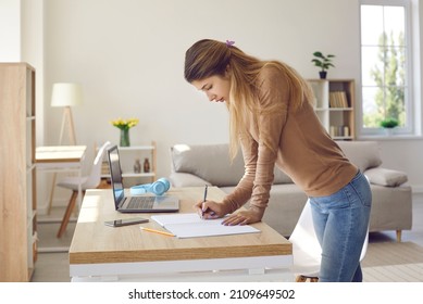 Young woman taking notes. Candid shot of college student standing by desk with paper, cellphone and laptop at home, doing homework, holding pen, looking at her notebook and thinking of what to write - Shutterstock ID 2109649502