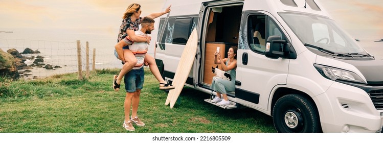 Young woman taking a photo with her cell phone of her friends having fun piggybacking next to their camper van during a trip - Powered by Shutterstock