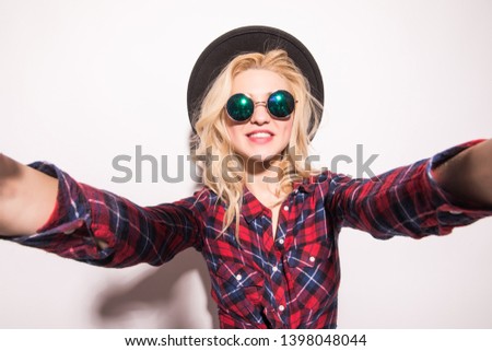 Young woman taking picture self portrait on smartphone wearing fashion black hat red checkered shirt over white background