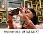 Young woman taking pictures of paintings in art museum with smartphone