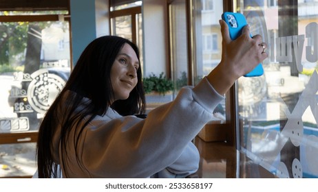 Young woman taking a selfie by a large window in a cozy café during daylight hours, enjoying a relaxed moment - Powered by Shutterstock