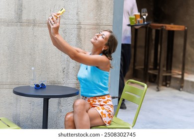 Young woman taking a selfie while sitting at a table in a colorful outfit during summer at a cozy outdoor café - Powered by Shutterstock