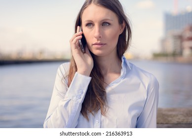 Young Woman Talking On Her Mobile Phone Listening To The Conversation With A Serious Expression As She Stands Outdoors Against A River Backdrop