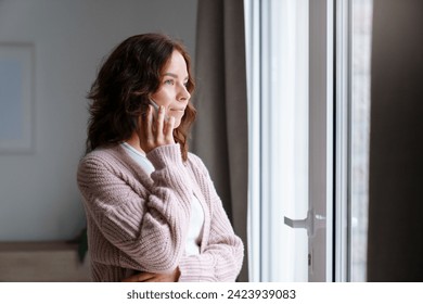 Young woman talking on mobile phone at home, looking through window. Tensed girl holding cell phone and calling, waiting. People connection, communication. Lifestyle moment - Powered by Shutterstock