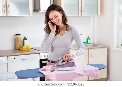 Happy Woman Ironing Cloth With Electric Iron In Kitchen, Stock