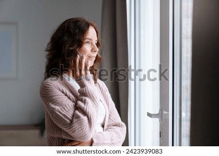 Young woman talking on mobile phone at home, looking through window. Tensed girl holding cell phone and calling, waiting. People connection, communication. Lifestyle moment