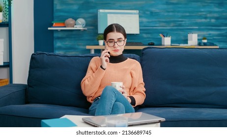 Young Woman Talking On Phone Call About School Courses, Using Smartphone To Discuss About Homework Assignment. College Student Sitting On Couch In Living Room And Chatting On Mobile Phone.