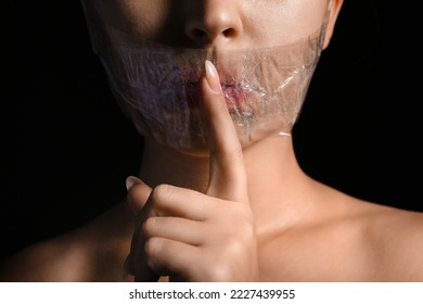 Young woman with taped mouth showing silence gesture on dark background, closeup. Censorship concept