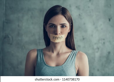 Young Woman With Taped Mouth With Word Food. Diet Concept. Weight Loss Problem. Starving Young Woman. Taped Mouth. Tape With Word. Hungry Woman. Healthy Lifestyle Concept. Gray Shirt.