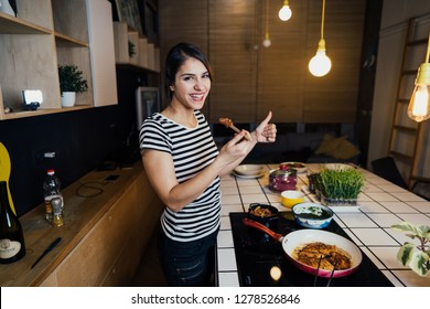 Young woman tasting a healthy meal in home kitchen.Making dinner on kitchen island standing by induction hob.Preparing fresh vegetables,enjoying spice aromas.Eating in.Passion for cooking.Dieting
