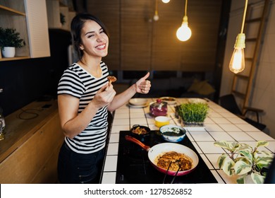 Young Woman Tasting A Healthy Meal In Home Kitchen.Making Dinner On Kitchen Island Standing By Induction Hob.Preparing Fresh Vegetables,enjoying Spice Aromas.Eating In.Passion For Cooking.Dieting