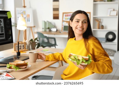 Young woman with tasty sandwich and cup of coffee in office - Shutterstock ID 2376265695