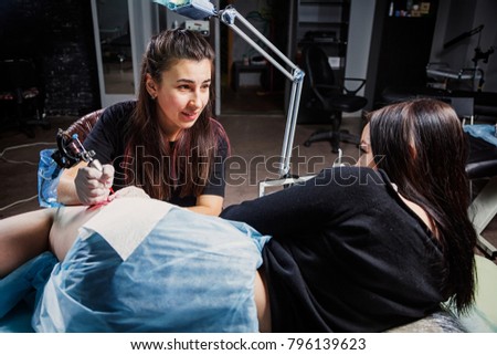 A young woman tattoo artist doing a tattoo with a tattoo machine under the bright light of a lamp on a beautiful woman with whom she talks in a dark workshop