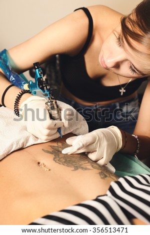 Young woman tattooer showing process of making a tattoo, black roses design on a stomach.