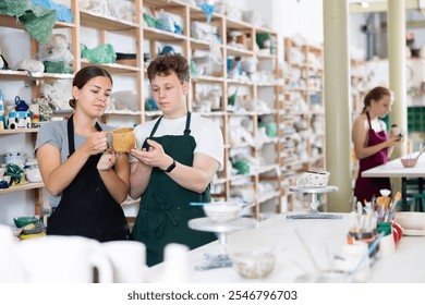 Young woman teacher in apron examines ceramic cup made by teenage boy student - Powered by Shutterstock